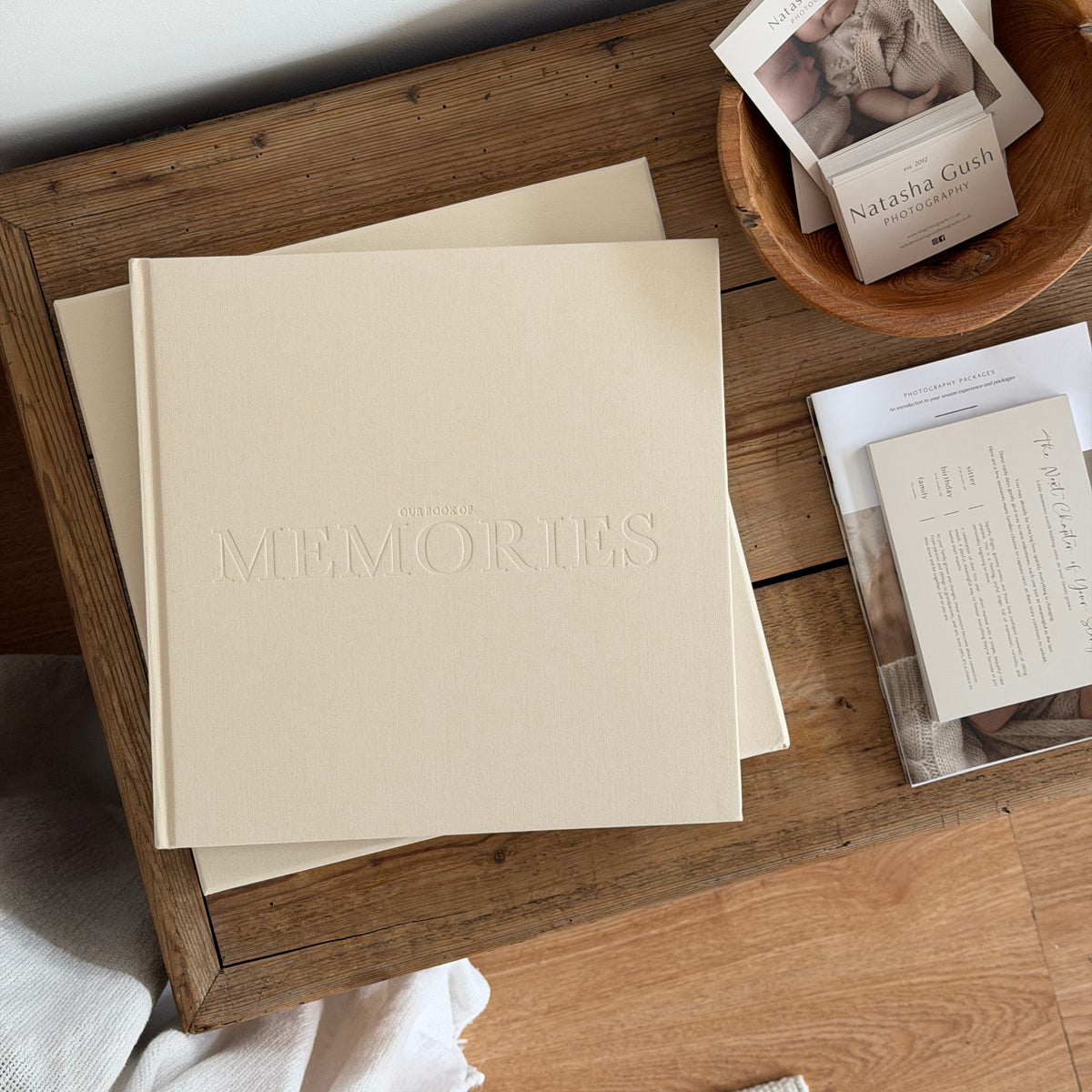 A cream-colored hardcover photo album with the word 'MEMORIES' embossed on the front, styled on a rustic wooden tray alongside other paper goods in a home setting.