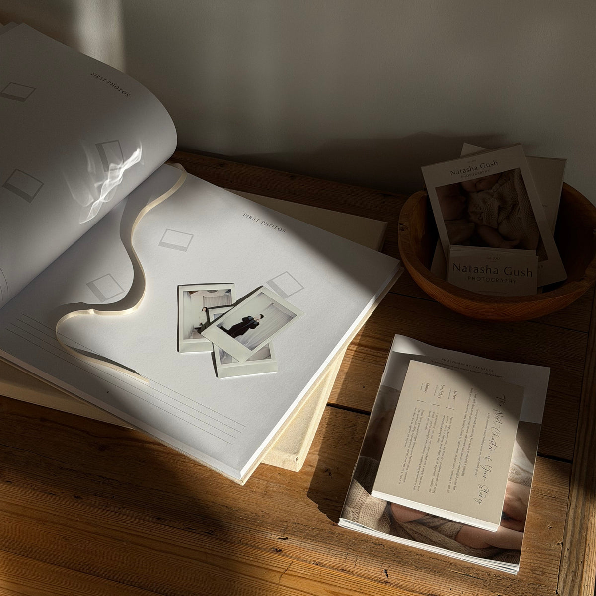 An open white photo album or book lies on a wooden table in warm sunlight, with a few small instant photographs placed on the pages. A small bowl of cards or prints sits nearby on the table against a neutral wall.