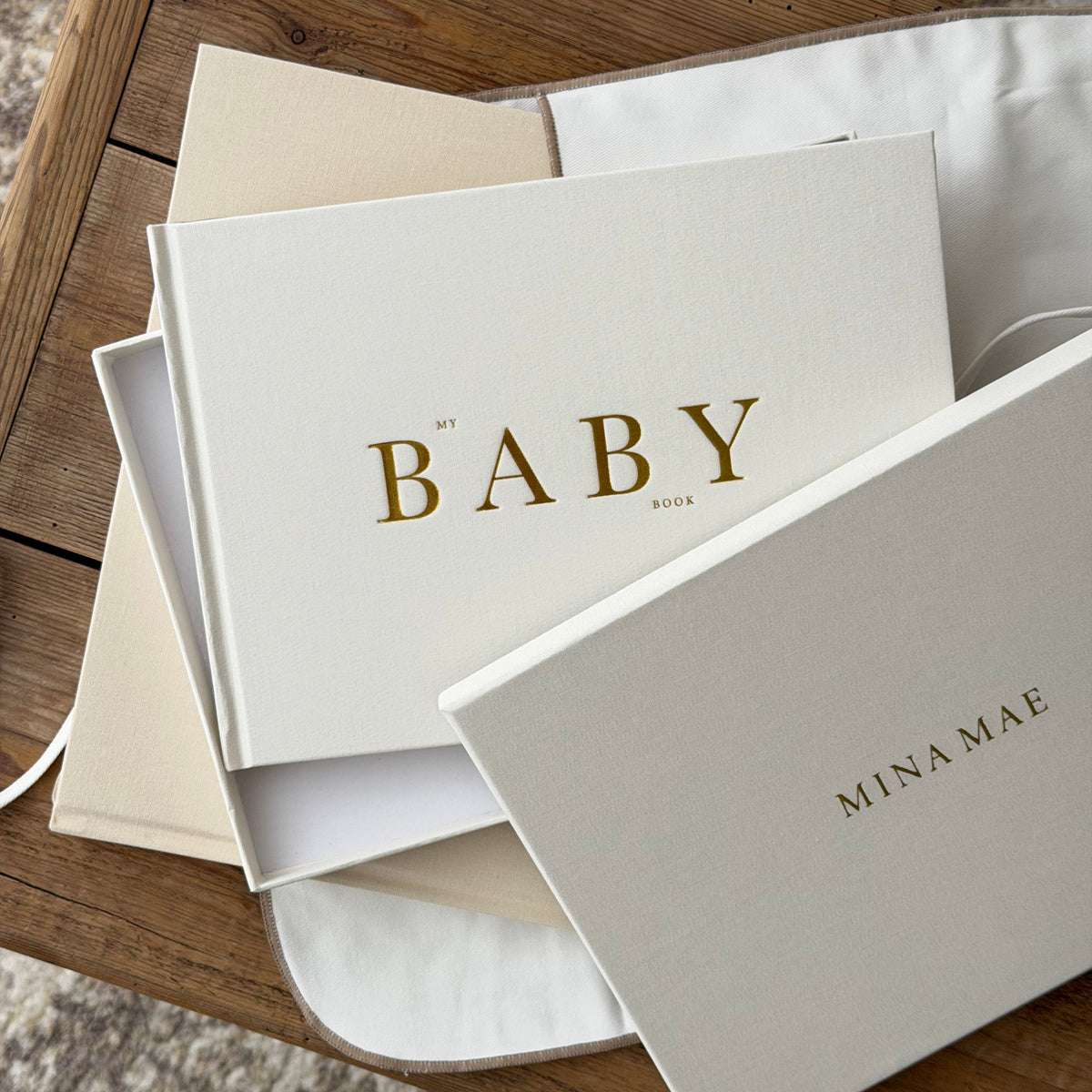 A stack of elegant baby keepsake boxes and envelopes in warm cream and beige tones, featuring gold foil lettering, arranged on a rustic wooden table.