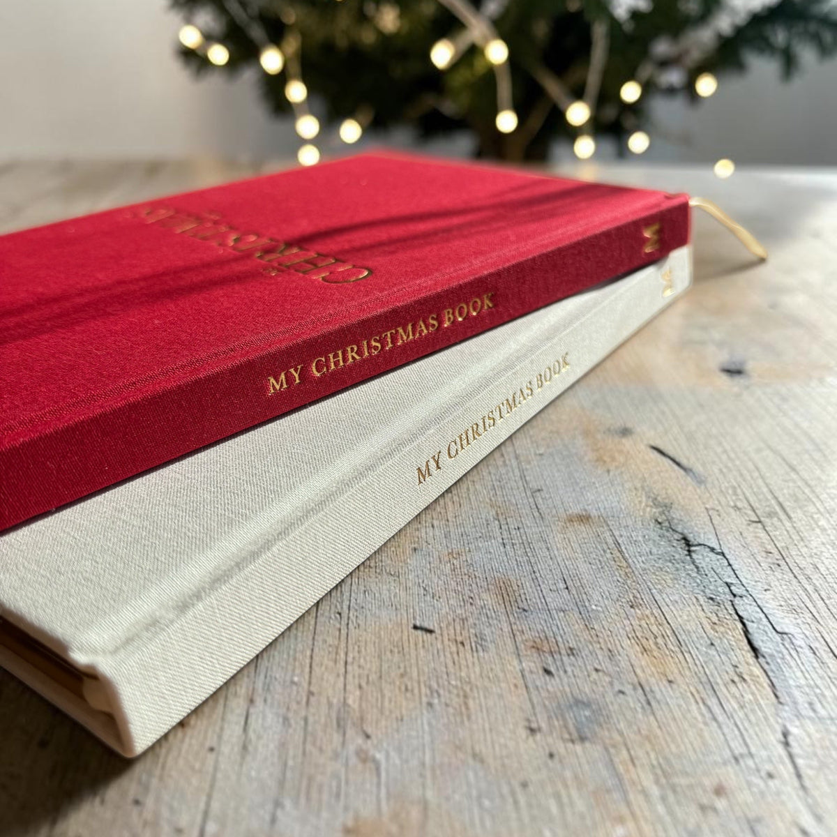 Two hardcover books titled 'My Christmas Book' stacked on a rustic wooden table, with one book in deep red velvet and the other in cream linen. A blurred Christmas tree with warm lights is in the background.